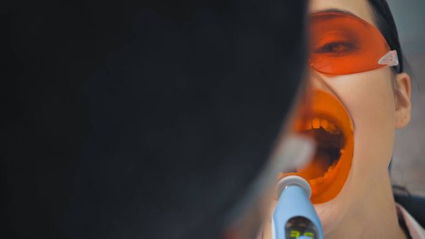 καλλιεργημένη άποψη της γυναίκας σε γυαλιά κοντά στον οδοντίατρο με υπεριώδη λάμπα, θολή πρώτο πλάνο - Φωτογραφία, εικόνα