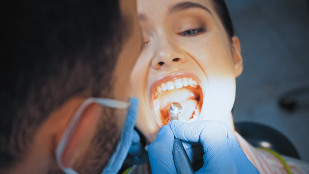 dentista em máscara médica usando broca dental durante o tratamento dos dentes da mulher, foreground borrado - Foto, Imagem