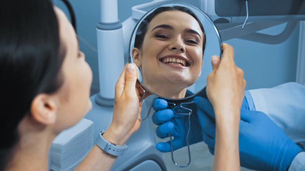 Frau mit perfekten Zähnen blickt in Spiegel neben Zahnarzt in Zahnklinik, verschwommener Vordergrund - Foto, Bild