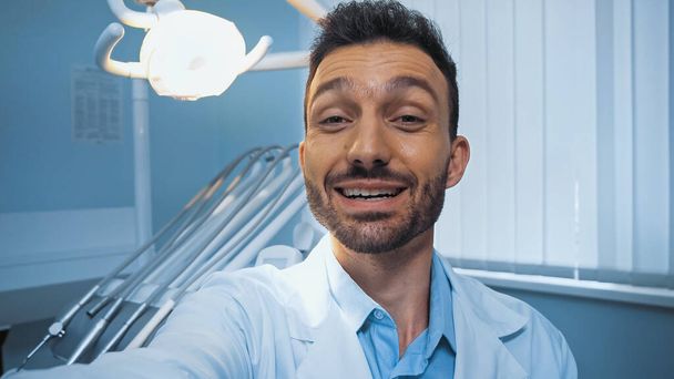 весёлый стоматолог, смотрящий в камеру возле лампы и стоматологического оборудования на размытом фоне - Фото, изображение