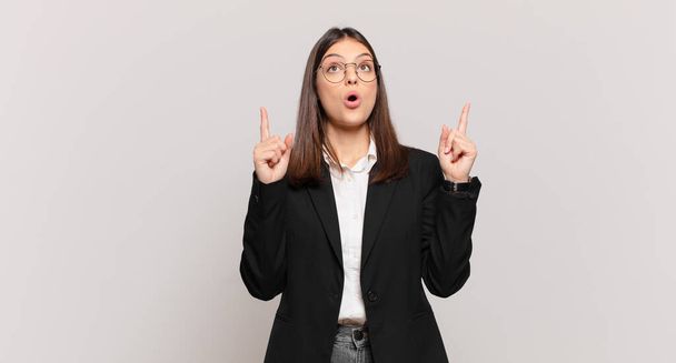 young business woman looking shocked, amazed and open mouthed, pointing upwards with both hands to copy space - 写真・画像
