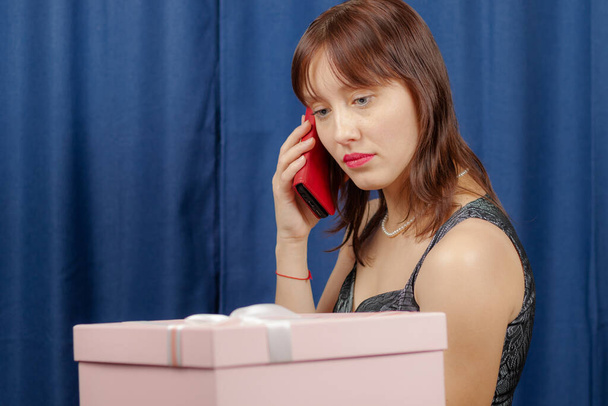 Şirin, kahverengi saçlı, elinde bir hediye olan pembe bir kutu tutan ve mavi arka planda telefonla konuşan bir kız. - Fotoğraf, Görsel