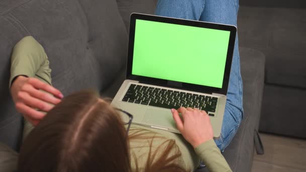 Jolie jeune femme caucasienne utilisant un ordinateur portable avec écran vert, couchée sur le canapé à la maison. Chromakey.  - Séquence, vidéo