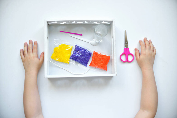 Flat lay kids pens are lying on a white table granules. Paraffin montessori material in three colors: yellow, orange, purple, scissors, candle cup, wick. - Photo, Image