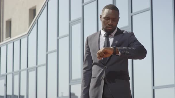 Low-angle Medium Shot mit Slowmo eines selbstbewussten afroamerikanischen Geschäftsmannes im Anzug, der auf seine Armbanduhr blickt und dann an einem sonnigen Tag im Geschäftsviertel das Taxi erwischt - Filmmaterial, Video