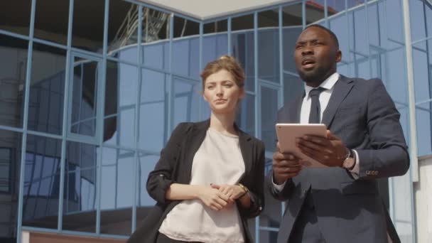 Low-angle Medium Shot mit Langsamkeit eines afrikanisch-amerikanischen Geschäftsmannes mit digitalem Tablet im Gespräch mit einer Geschäftspartnerin, die irgendwo in der Nähe eines modernen gläsernen Bürogebäudes steht - Filmmaterial, Video
