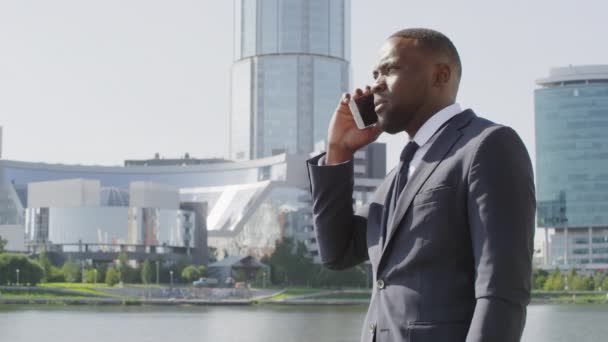 Plano mediano de bajo ángulo del empresario afroamericano confiado en traje que habla por teléfono mientras está parado en el distrito de negocios de la ciudad moderna en el día soleado - Metraje, vídeo