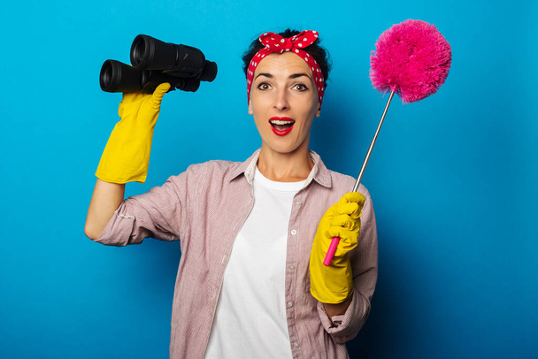 Surprised young woman in cleaning gloves holding binoculars and dust brush on blue background - Photo, Image