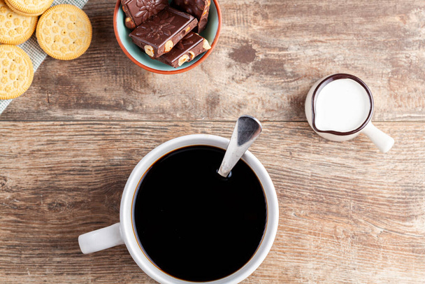 ένα τσάι ή ώρα καφέ έννοια με μπισκότα σάντουιτς, μπαρ από σοκολάτα αισθητική κεραμικά κούπα και πιάτα, καθώς και μια μίνι κανάτα κρέμα σε ξύλινο τραπέζι. - Φωτογραφία, εικόνα