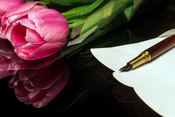 Stílusos toll egy képeslapon gratulációkhoz és virágok átlátszó csomagolásban a háttérben. Ötlet egy évfordulós vagy ünnepi üdvözlőlevél megírására - Fotó, kép