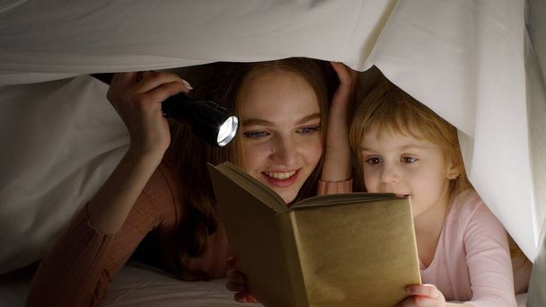 Joven madre leyendo buenas noches cuento de hadas a la hija del niño bajo manta nórdica en la habitación de noche - Foto, Imagen
