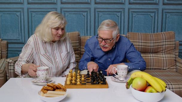 Rentnerehepaar trinkt Tee, spielt Schach in modernem Wohnzimmer Wohnzimmer zusammen - Foto, Bild