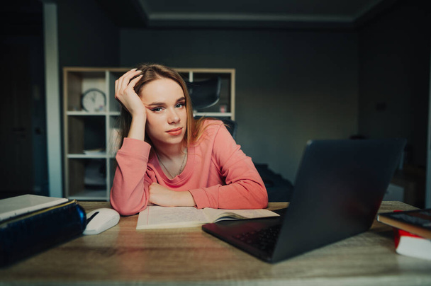 Estudante feminina perturbada adormece em casa em uma mesa durante aulas de internet on-line em um laptop, olha para a câmera com um rosto triste e fica atrás do local de trabalho. - Foto, Imagem
