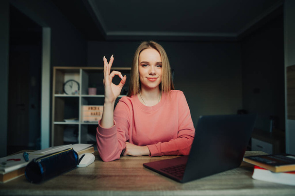 Linda mujer joven en un suéter rosa se sienta en casa en su escritorio y muestra gesto OK, mirando a la cámara. Aprender en línea es bueno. - Foto, imagen