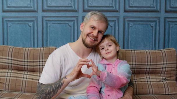 Father and child daughter kid in pajamas sit on couch smiling, making sign of shape heart near chest - Photo, Image