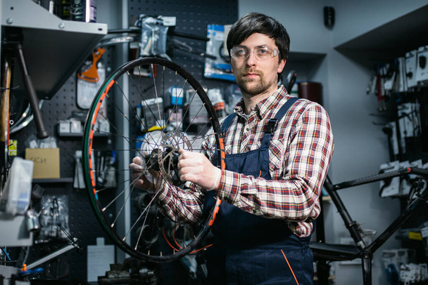 Mecánico masculino que trabaja en taller de reparación de bicicletas, mecánico que repara la bicicleta usando la herramienta especial, usando guantes protectores. Joven y atractivo militar arreglando la rueda de bicicleta del cliente en su propio taller. - Foto, imagen