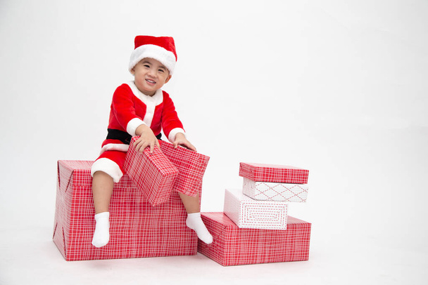 Menino asiático feliz com Papai Noel terno segurando caixa de presente e sentado na caixa de presente vermelho isolado no fundo branco - Foto, Imagem