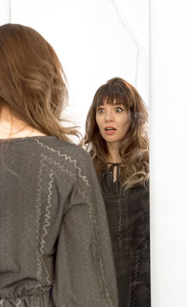 感情的に美しい若いブルネットが鏡に立って顔を作る - 写真・画像