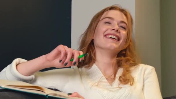 Affascinante donna zenzero con lentiggini sorride e ride mentre fa i compiti usando una penna e un copybook sul divano - Filmati, video