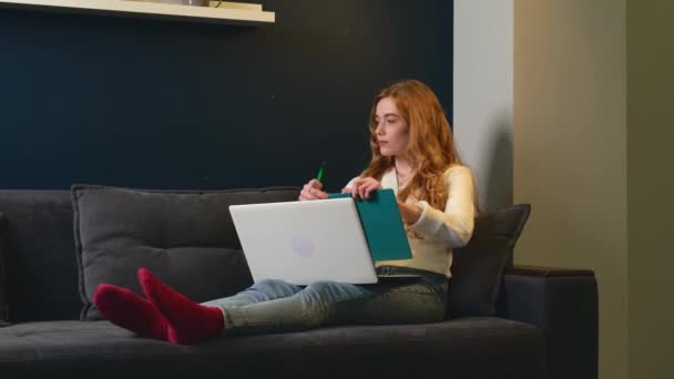 Donna caucasica con i capelli rossi e lentiggini sta scrivendo qualcosa mentre utilizza un computer portatile sdraiato su un divano - Filmati, video