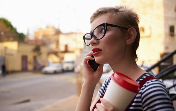 mulher com cabelo curto falando ao telefone na rua e uma xícara de café na mão - Foto, Imagem