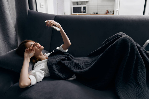 μια γυναίκα ξαπλώνει σε έναν καναπέ σε ένα διαμέρισμα με ένα κινητό τηλέφωνο στο χέρι της - Φωτογραφία, εικόνα