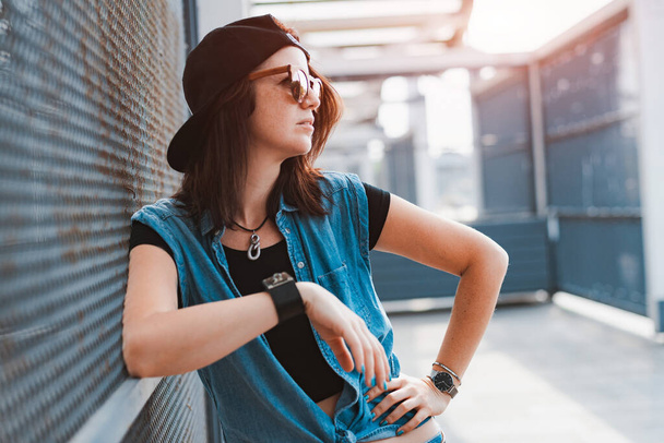 jeune fille hipster élégant dans une image audacieuse dans une casquette et des lunettes de soleil pose dans la ville dans les rayons du coucher du soleil sur un fond urbain. - Photo, image