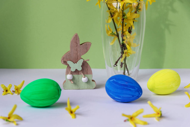 Painted eggs, wooden toy bunny and forsythia yellow flowers in a glass. Easter concept. - Photo, Image