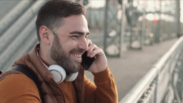 Médio close-up de feliz homem caucasiano de olhos azuis em pé na ponte, sorrindo, falando no telefone celular no dia ensolarado - Filmagem, Vídeo