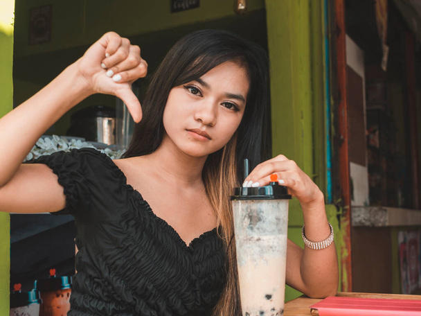 Egy depressziós, fiatal ázsiai nő lenyom egy hüvelykujjat, miközben tejet szürcsöl. Lehetséges negatív felülvizsgálat vagy elutasítás. - Fotó, kép