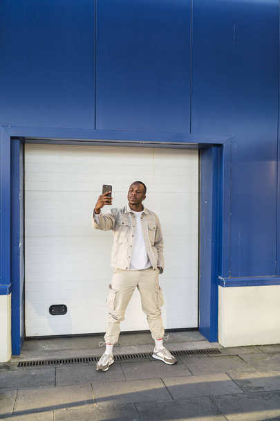 A vertical shot of a black male taking a selfie outdoors near a blue wall - Fotoğraf, Görsel