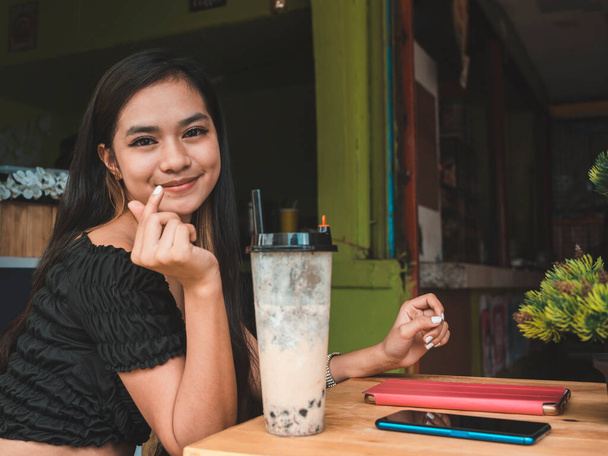 Симпатичный и привлекательный молодой филиппинки женщина дает знак сердца палец, потягивая чай молока. - Фото, изображение