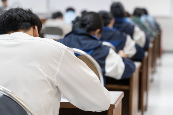Rear view of the teenage college students sit on lecture chairs do final examination and write on examination paper answer sheets in the classroom. University students in uniform in classroom. - Photo, Image