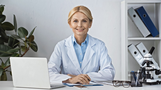 счастливый доктор улыбается в камеру, сидя рядом с ноутбуком на рабочем месте - Фото, изображение
