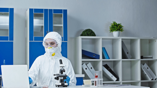 учёный в защитном костюме работает рядом с ноутбуком и микроскопом в лаборатории - Фото, изображение