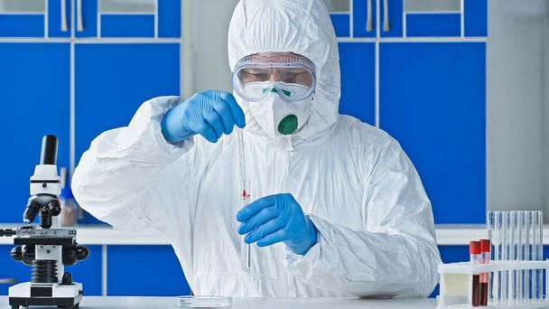 científico en traje de materiales peligrosos que sostiene el tubo de ensayo y el gotero mientras trabaja cerca del microscopio - Foto, imagen