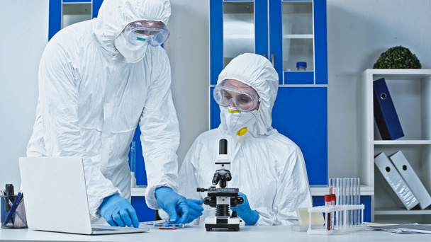 científico en traje de materiales peligrosos que muestra la muestra de sangre a su colega cerca del microscopio - Foto, Imagen