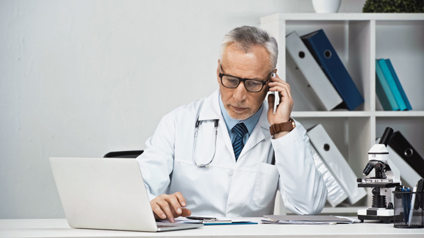 serieuze arts praten op smartphone in de buurt van laptop in het ziekenhuis - Foto, afbeelding