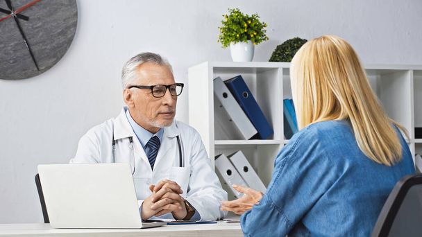ξανθιά γυναίκα που μιλάει με προσεκτικό γιατρό κατά τη διάρκεια του ραντεβού στην κλινική - Φωτογραφία, εικόνα