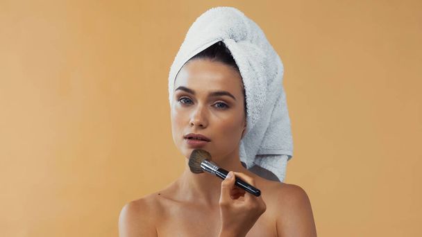 bonita mujer con toalla en la cabeza aplicando polvo facial con cepillo cosmético aislado en beige - Foto, Imagen