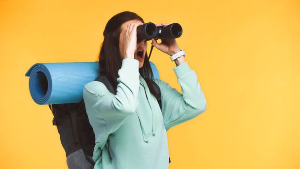 Excited traveler with backpack looking through binoculars isolated on yellow - Photo, Image