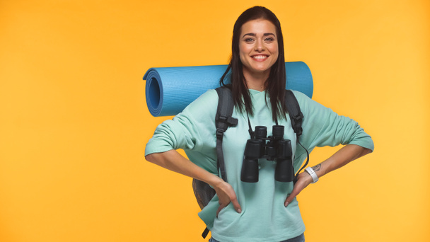 счастливый путешественник с рюкзаком, фитнес-мат и бинокль стоя с руками на бедрах изолированы на желтый - Фото, изображение