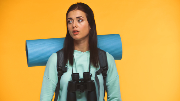 потерянный путешественник с рюкзаком, фитнес-мат и бинокль глядя изолированы на желтый - Фото, изображение