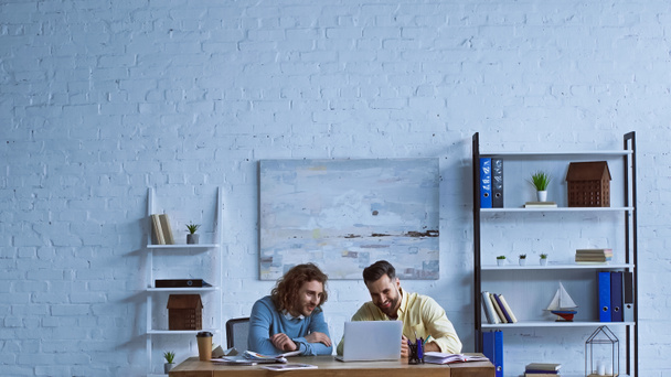 веселые бизнесмены, работающие рядом с ноутбуком в современном офисе - Фото, изображение