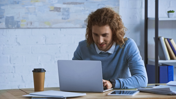 homem de negócios encaracolado digitando no laptop perto de café para ir ao local de trabalho - Foto, Imagem