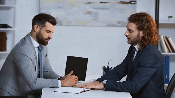 jonge zakenmannen in formele kleding praten in de buurt laptop in kantoor - Foto, afbeelding