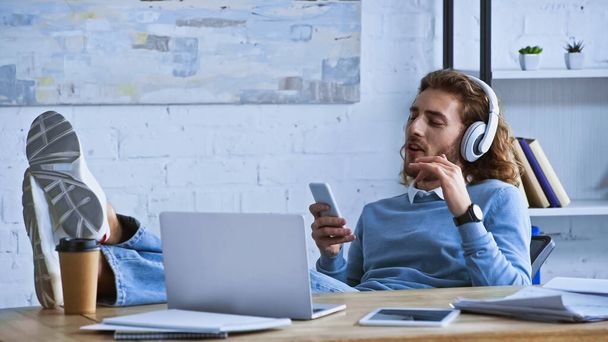 молодий бізнесмен слухає музику і спілкується на смартфоні, розслабляючись в офісі
 - Фото, зображення