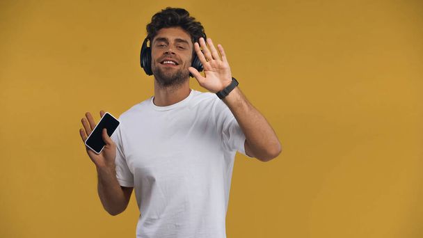 happy man in wireless headphones holding smartphone and dancing isolated on yellow - Photo, Image