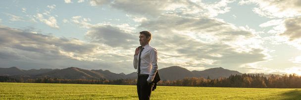 Jeune homme d'affaires confiant debout avec sa veste au-dessus de l'épaule dans une belle prairie verte, regardant au loin avec confiance. - Photo, image