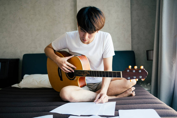 Young thai guitarist man seriously practice acoustic guitar on a bed, reading lyric paper notes. - Photo, Image